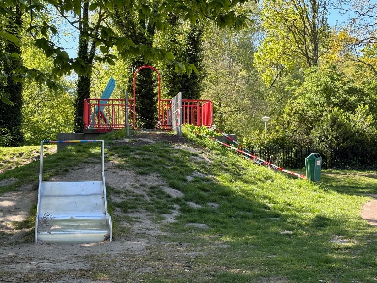 Wide metal slide built into a grass hill and red railing on the top with trees in the background