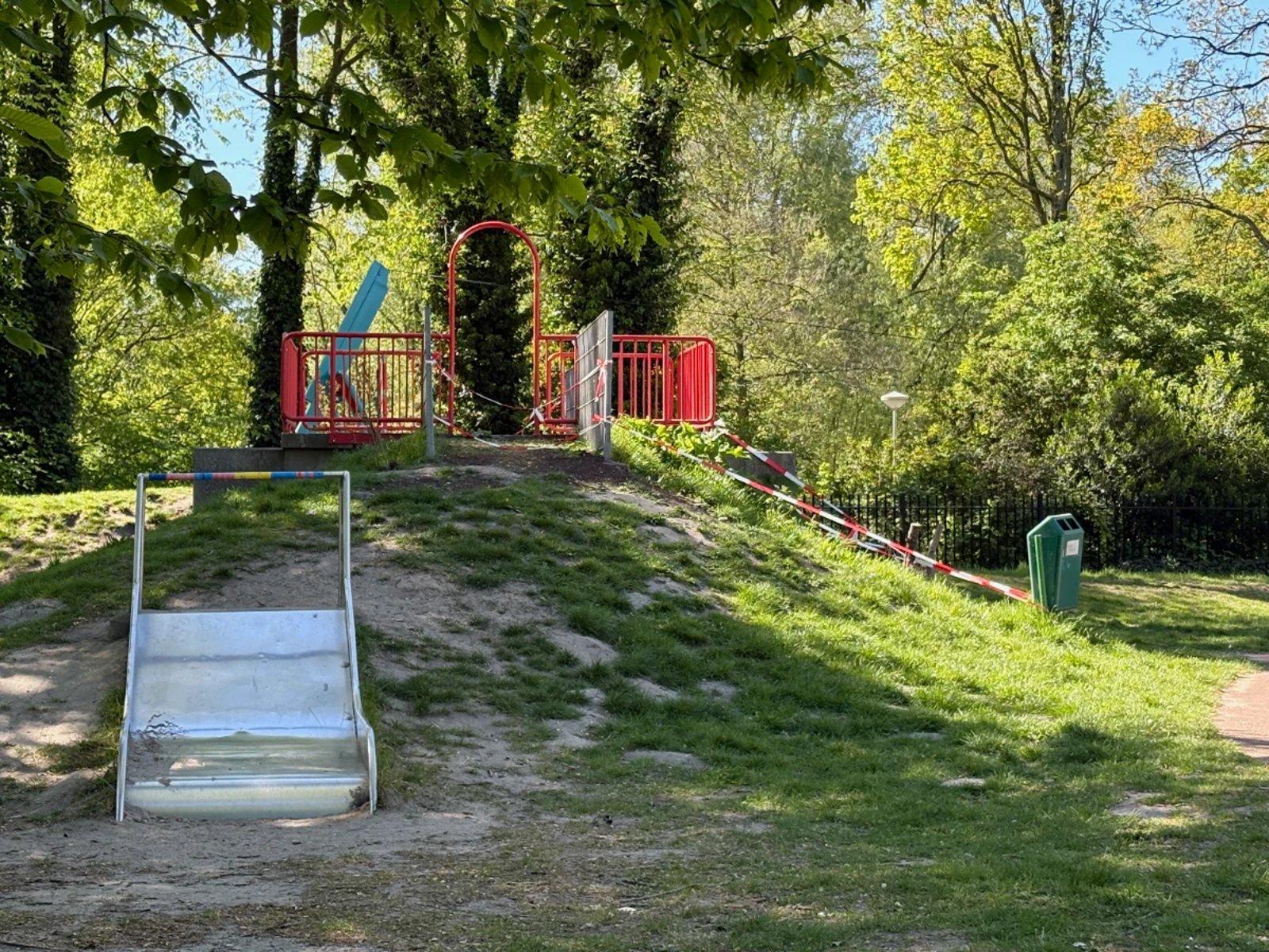 Wide metal slide built into a grass hill and red railing on the top with trees in the background