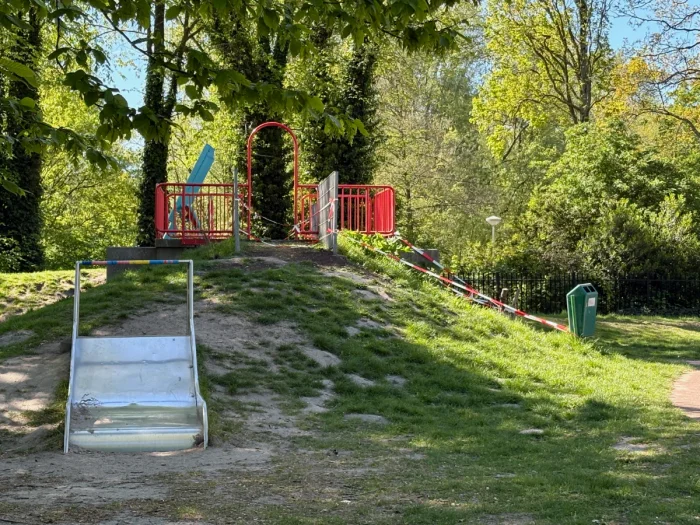 Wide metal slide built into a grass hill and red railing on the top with trees in the background