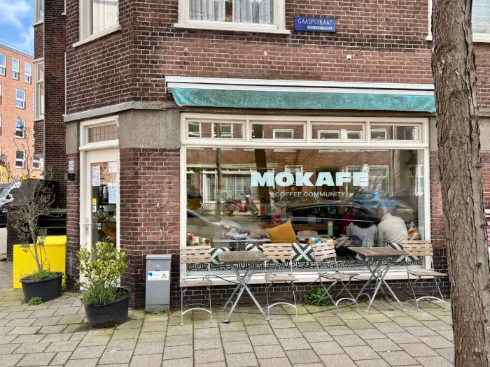 Corner coffeeshop with tables and chairs out front and a large window with MOKAFE text and people sitting inside