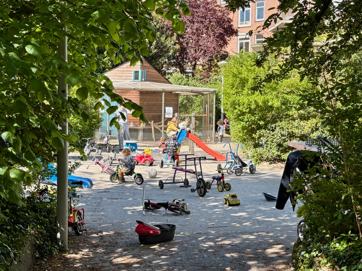 Shaded entrance to a children's playground with kids playing on bikes and a small slide and a chicken coop in the back