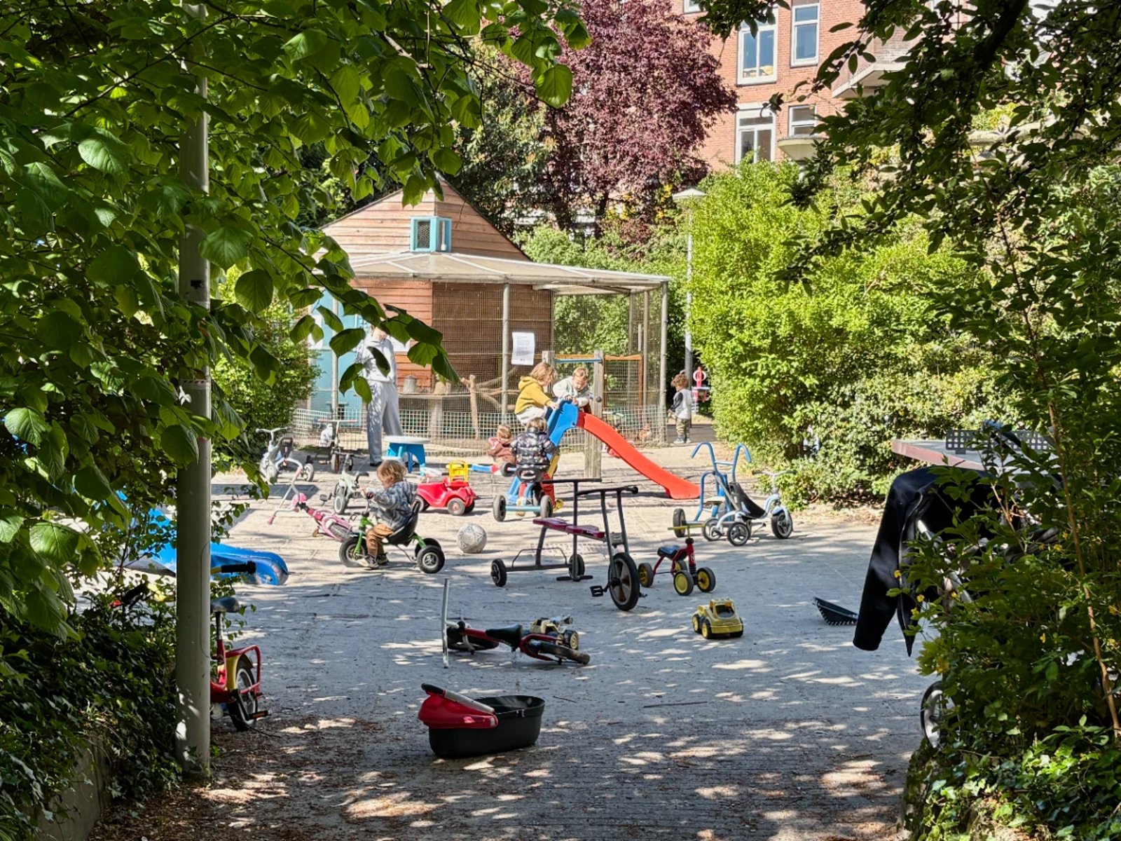 Shaded entrance to a children's playground with kids playing on bikes and a small slide and a chicken coop in the back