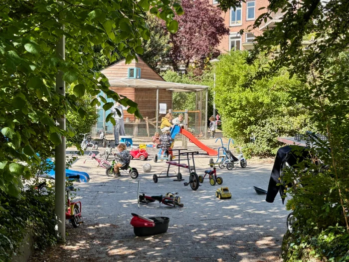 Shaded entrance to a children's playground with kids playing on bikes and a small slide and a chicken coop in the back