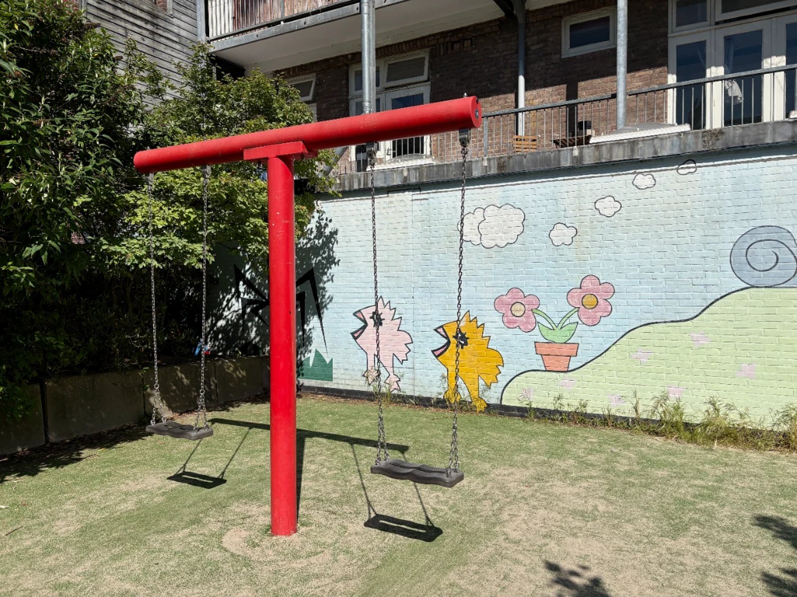 Two swings on a bright red swing set with a colorful mural behind showing chicks and flowers