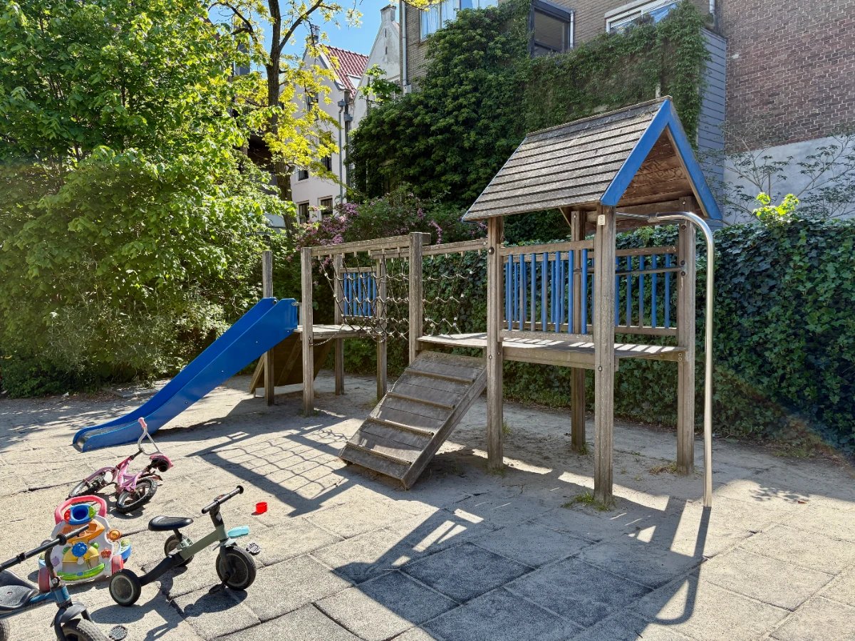Wooden play house structure with blue slide, rope bridge, and ramp with trees and buildings behind