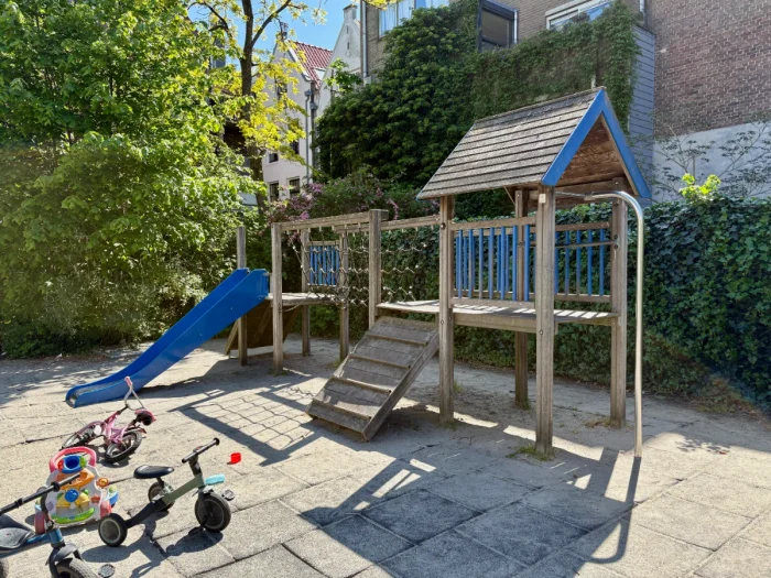 Wooden play house structure with blue slide, rope bridge, and ramp with trees and buildings behind