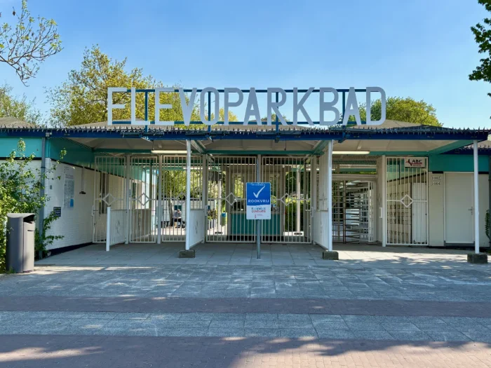 Gated entrance to public pool with white walls and gate, teal accents, and large FLEVOPARKBAD sign on roof