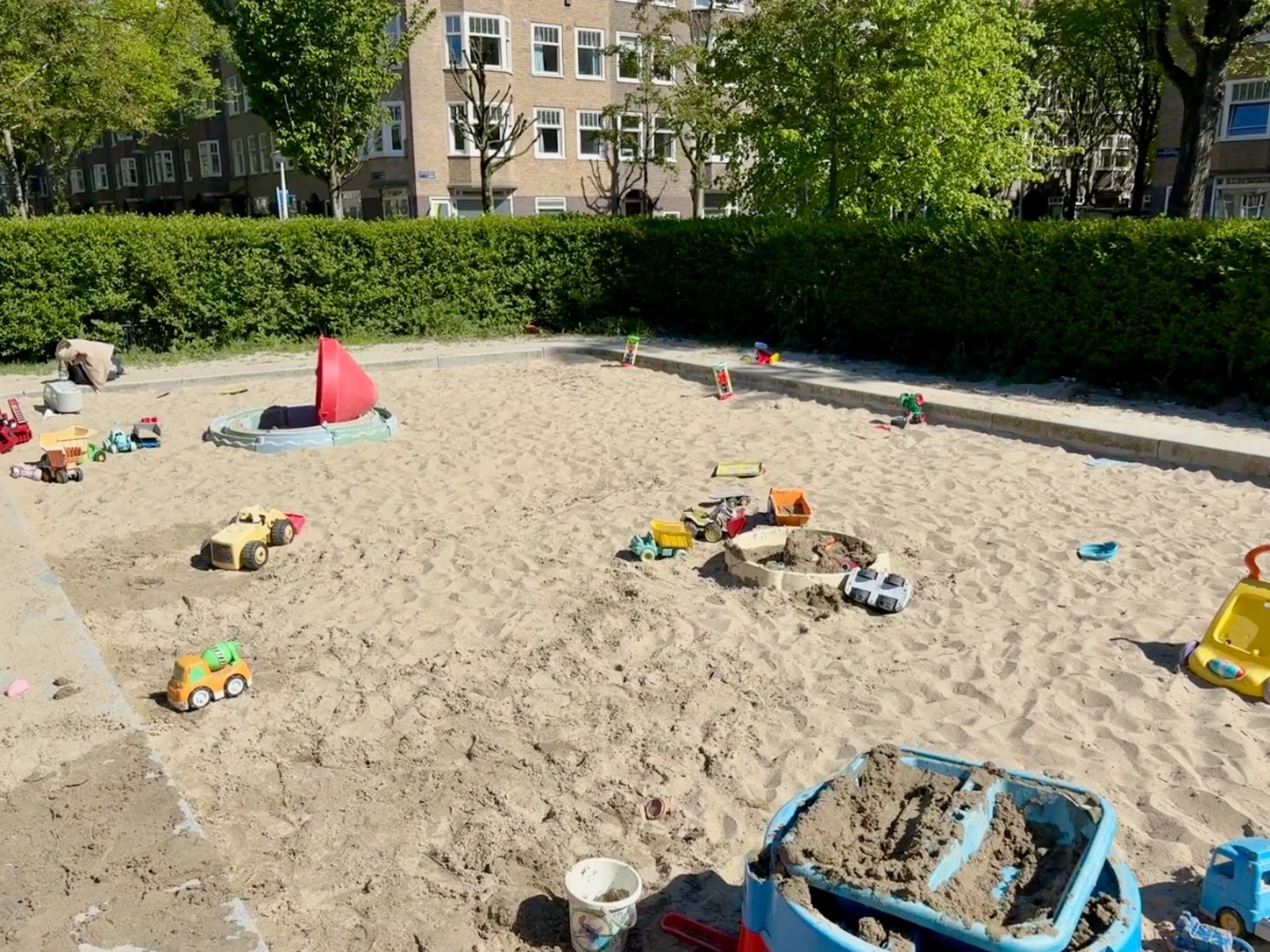 Large sandbox on sunny day with toys scattered about and residential buildings seen in the distance