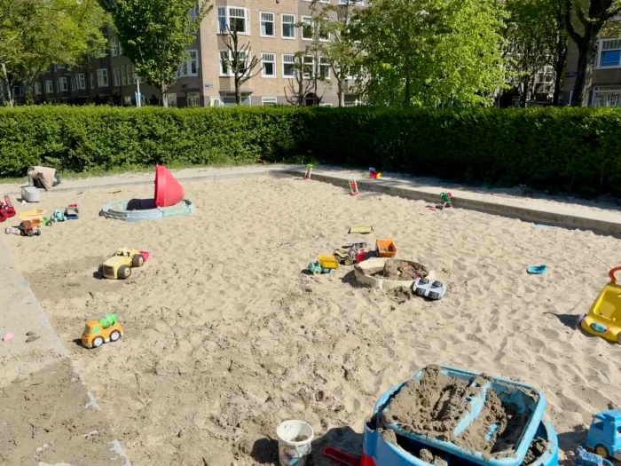 Large sandbox on sunny day with toys scattered about and residential buildings seen in the distance