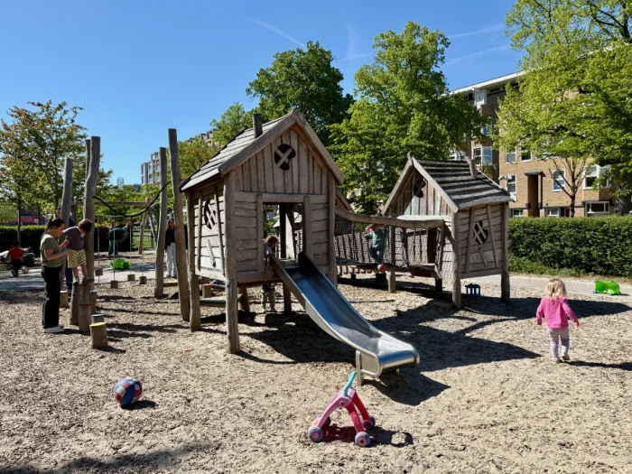 Kids and adults playing on wooden play house structure with slide, rope bridge, and sand