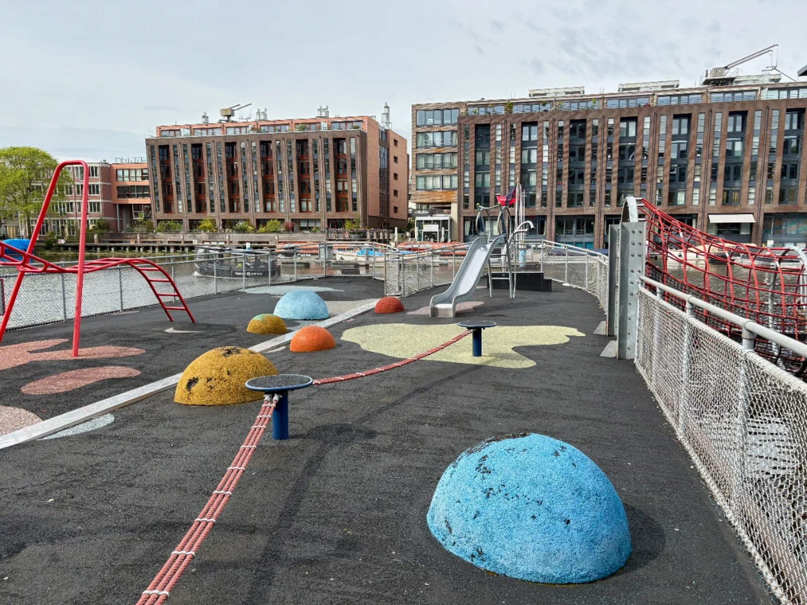 Barge playground on the water with red ladder, balance rope, slide, and blue, yellow, and orange sheres