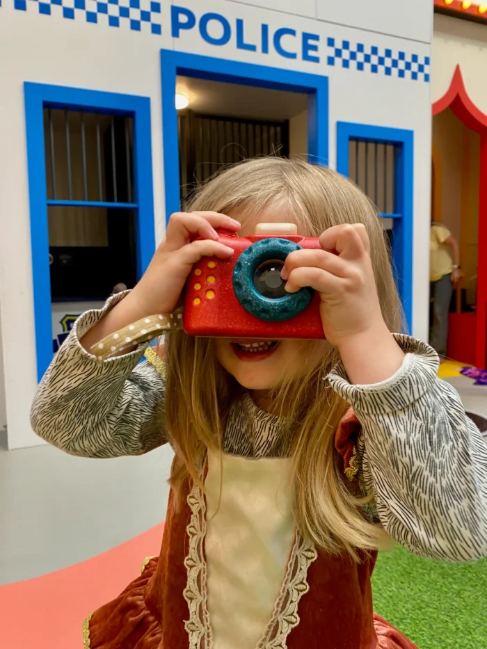 Girl taking a photo with a toy camera with a bright blue and white play police station behind