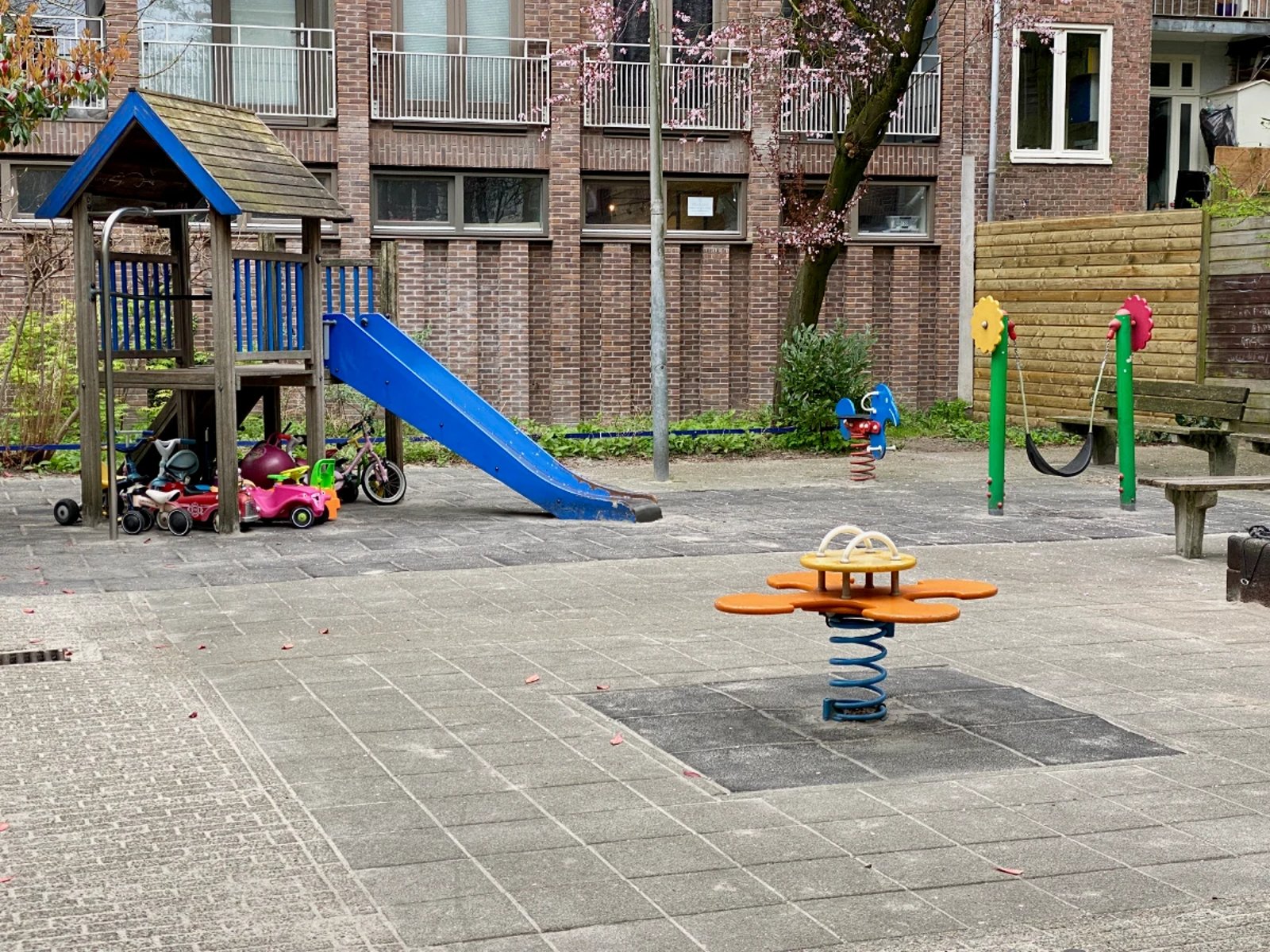 Enclosed playground with wooden house and blue slide, orange spring rocker, and green toddler swing with flowers