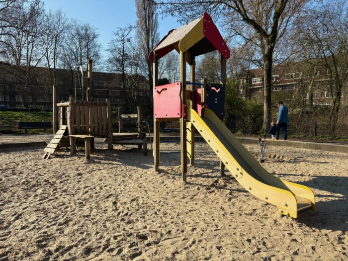 Outdoor sandy playground with red play house and yellow slide and small wooden climbing structure