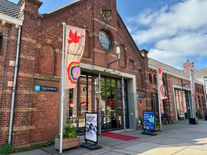 Historic brick building with large glass storefront, posters outside, and flags by the entrance with a rainbow and doves
