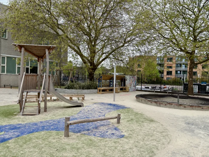 Small play house with slide, covered sandbox, and balance beam on sandy playground with large trees and buildings