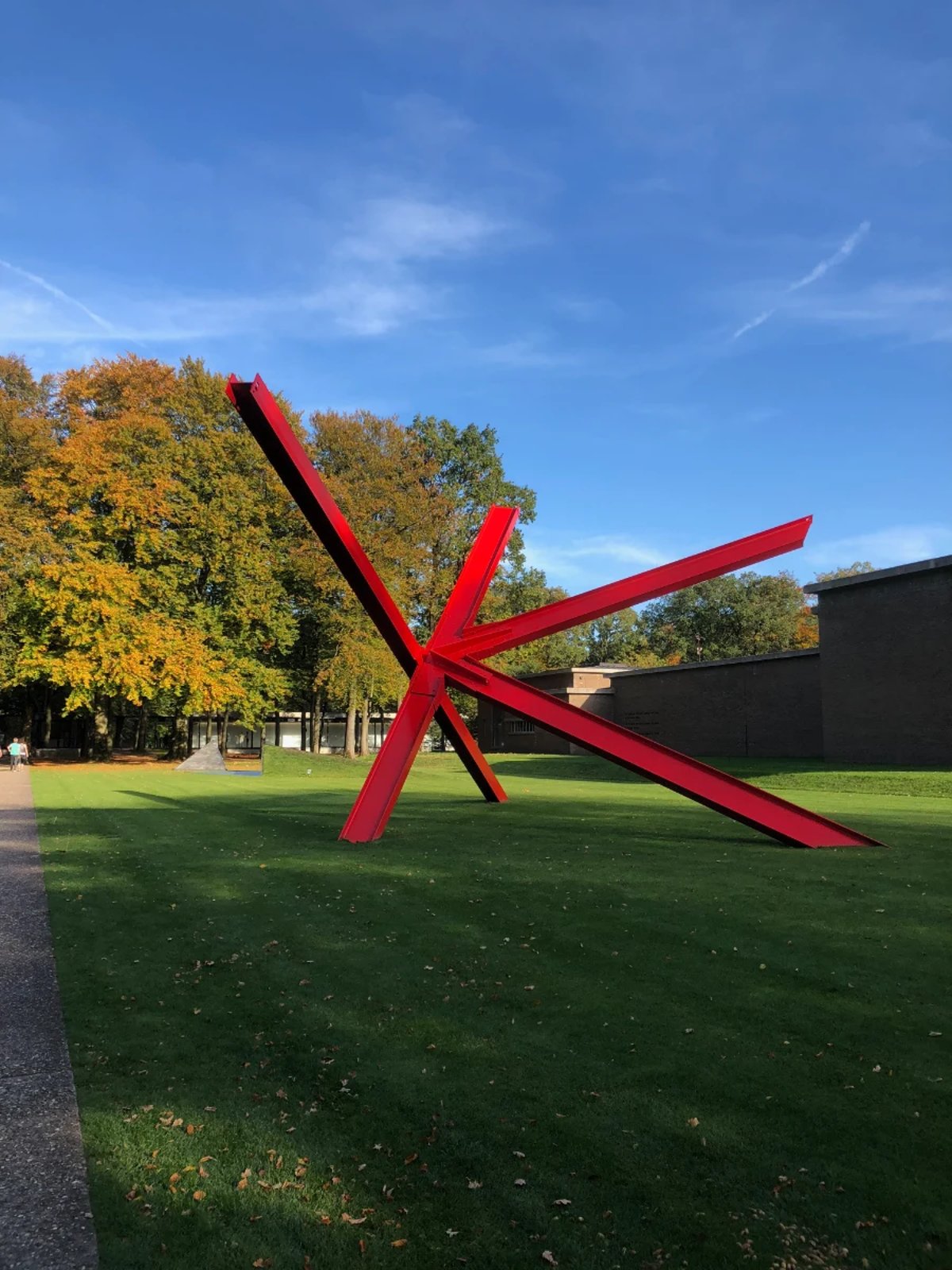 Large red outdoor sculpture on grassy lawn with autumn trees and blue skies in the background