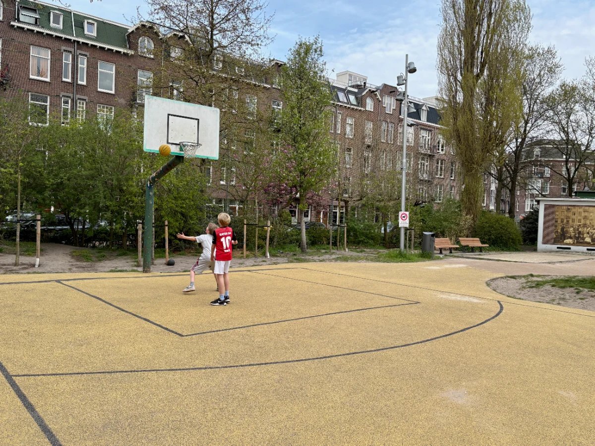 Two kids shoot at basketball hoop in a park with residential buildings in the distance