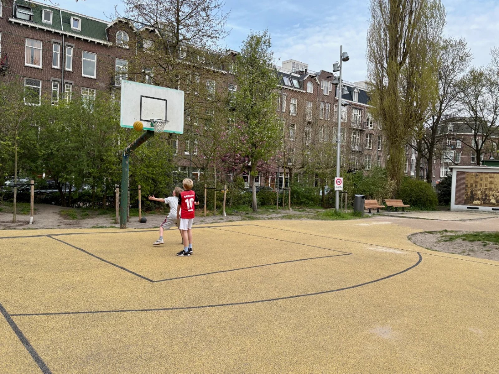 Two kids shoot at basketball hoop in a park with residential buildings in the distance