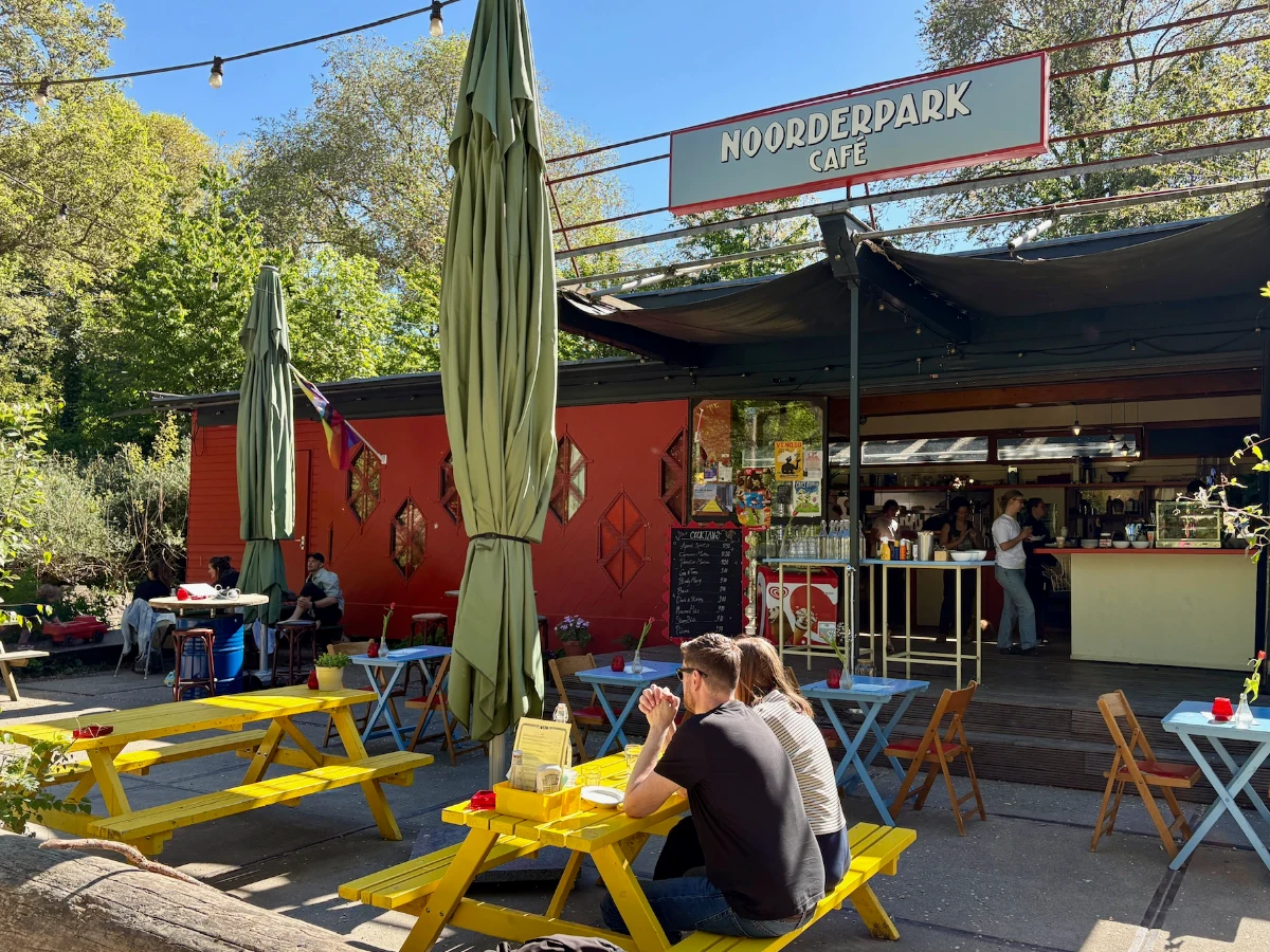 Red-painted cafe with yellow picnic tables, blue tables, and green sun umbrellas on the terrace and a large sign above the entrance