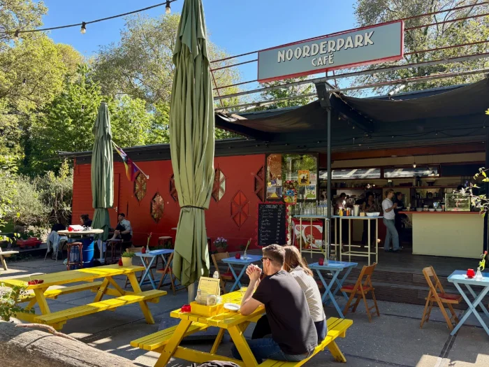 Red-painted cafe with yellow picnic tables, blue tables, and green sun umbrellas on the terrace and a large sign above the entrance