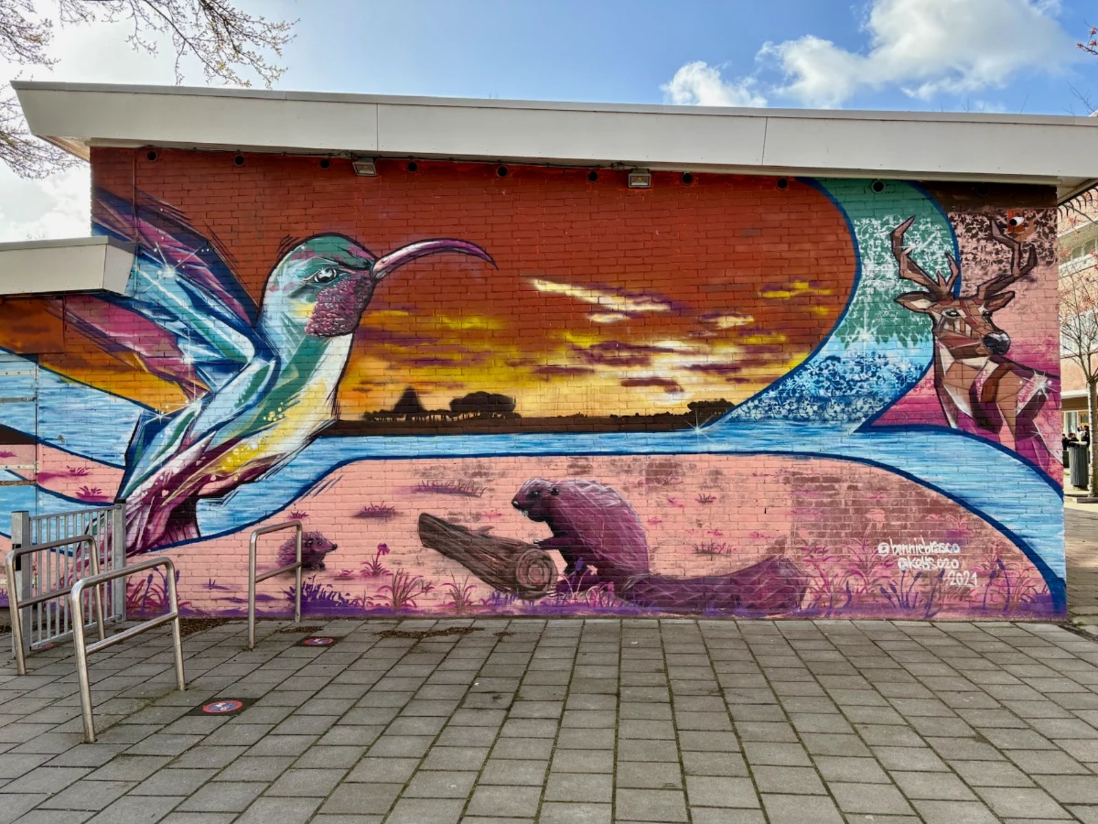 Mural on brick building of humming bird, beaver, and deer with sunset and viivid blue, red, and pink hues