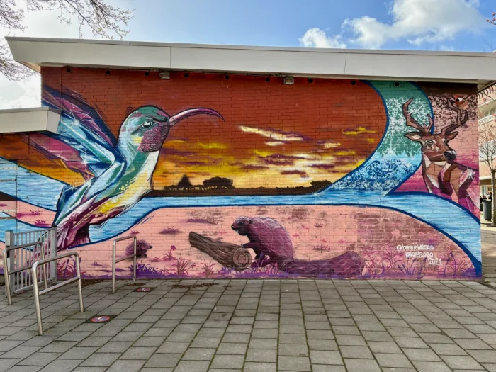 Mural on brick building of humming bird, beaver, and deer with sunset and viivid blue, red, and pink hues