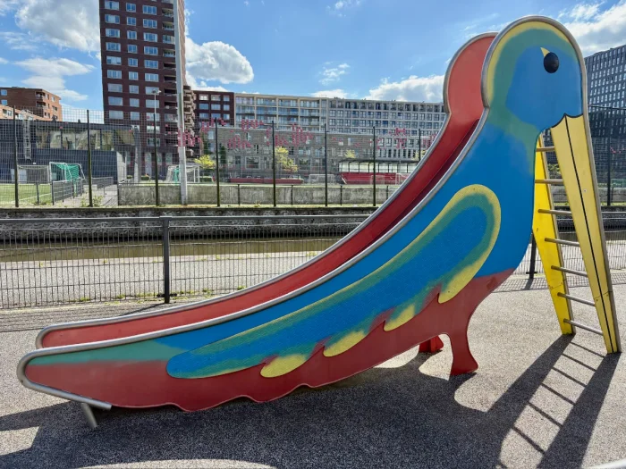 Blue, red, and yellow bird-shaped slide with sports fields behind