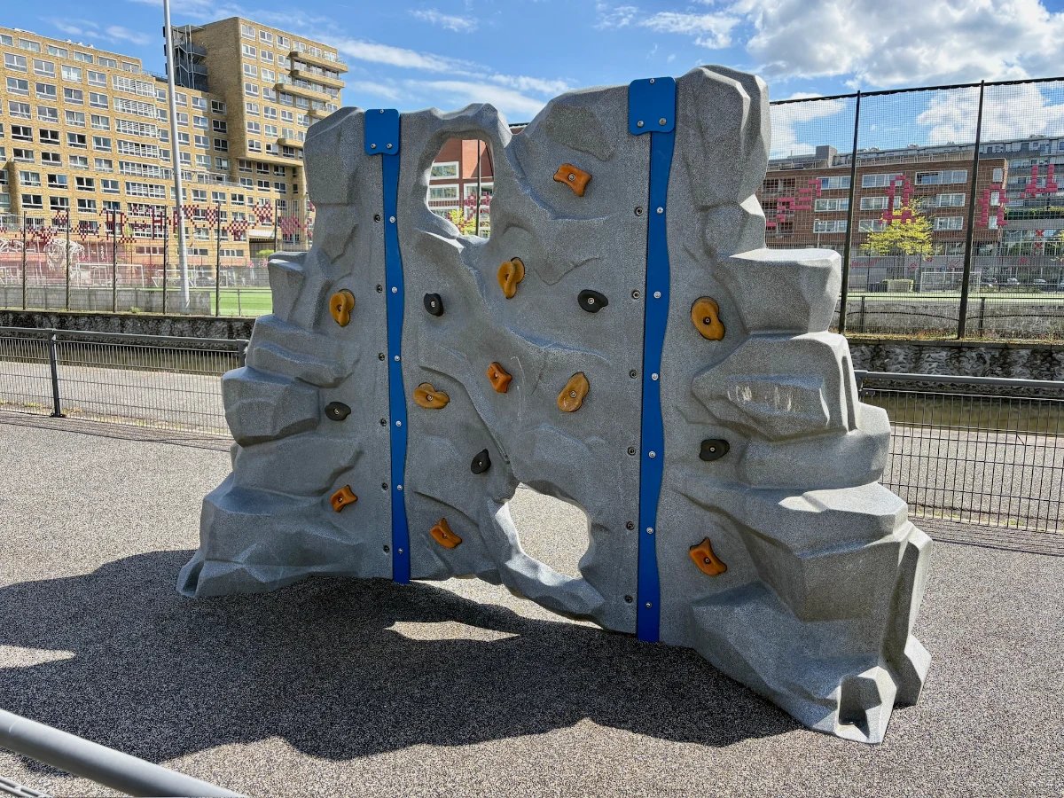 Grey climbing rock with blue and orange holds and sportsfields behind