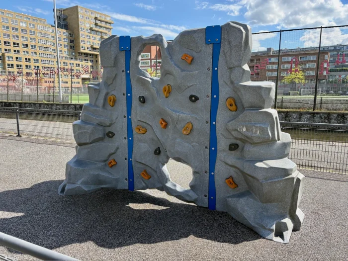 Grey climbing rock with blue and orange holds and sportsfields behind