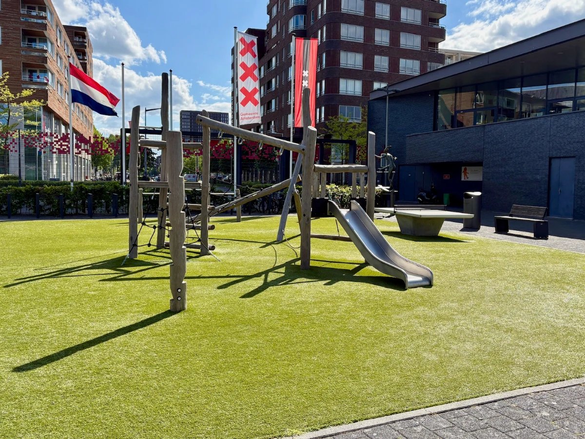 Wooden climbing structure with slide on sunny astroturf with banners and buildings behind