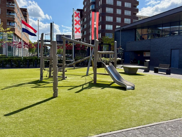 Wooden climbing structure with slide on sunny astroturf with banners and buildings behind
