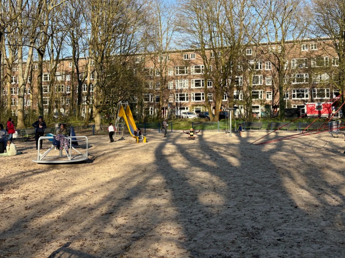 Parents and kids at playground with merry-go-round, yellow slide and residential building with trees behind and large shadow of a tree covering the ground