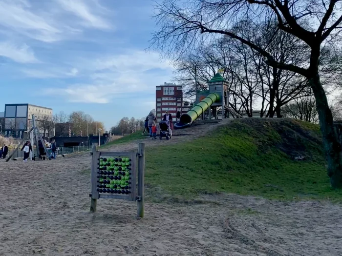 Large sandy play area with mature tree, green puzzle and big slide on top of a hill