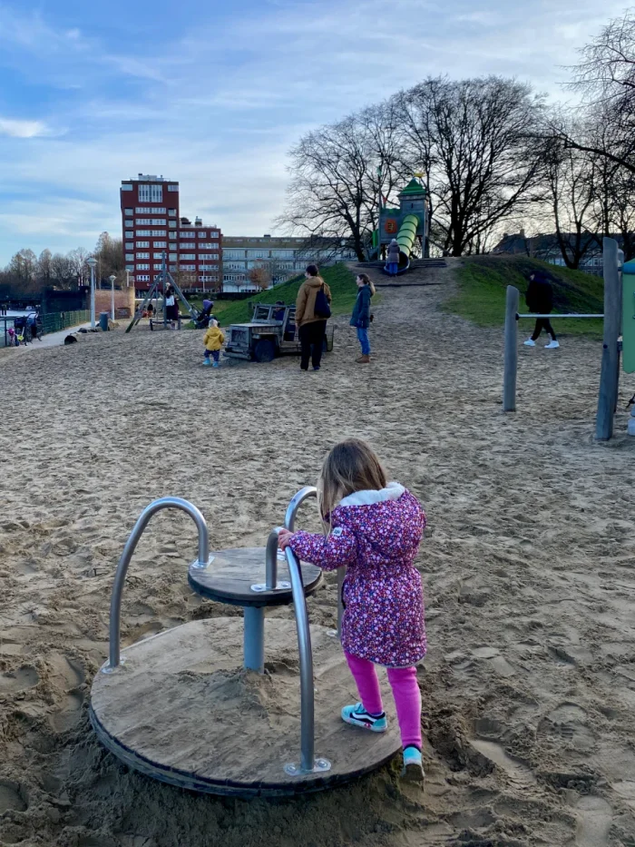 Girl in a pink coat on a merry-go-round with large sandy area and big slide on top of the hill