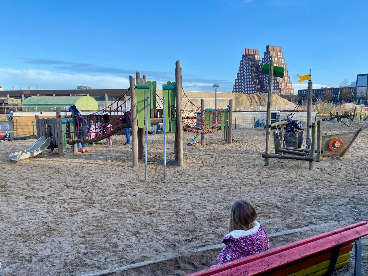 Rustic playground with wooden pirate ship and green climbing structure set on sand with modern pyramind-shaped residential building on the horizon