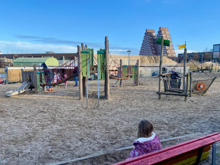 Rustic playground with wooden pirate ship and green climbing structure set on sand with modern pyramind-shaped residential building on the horizon