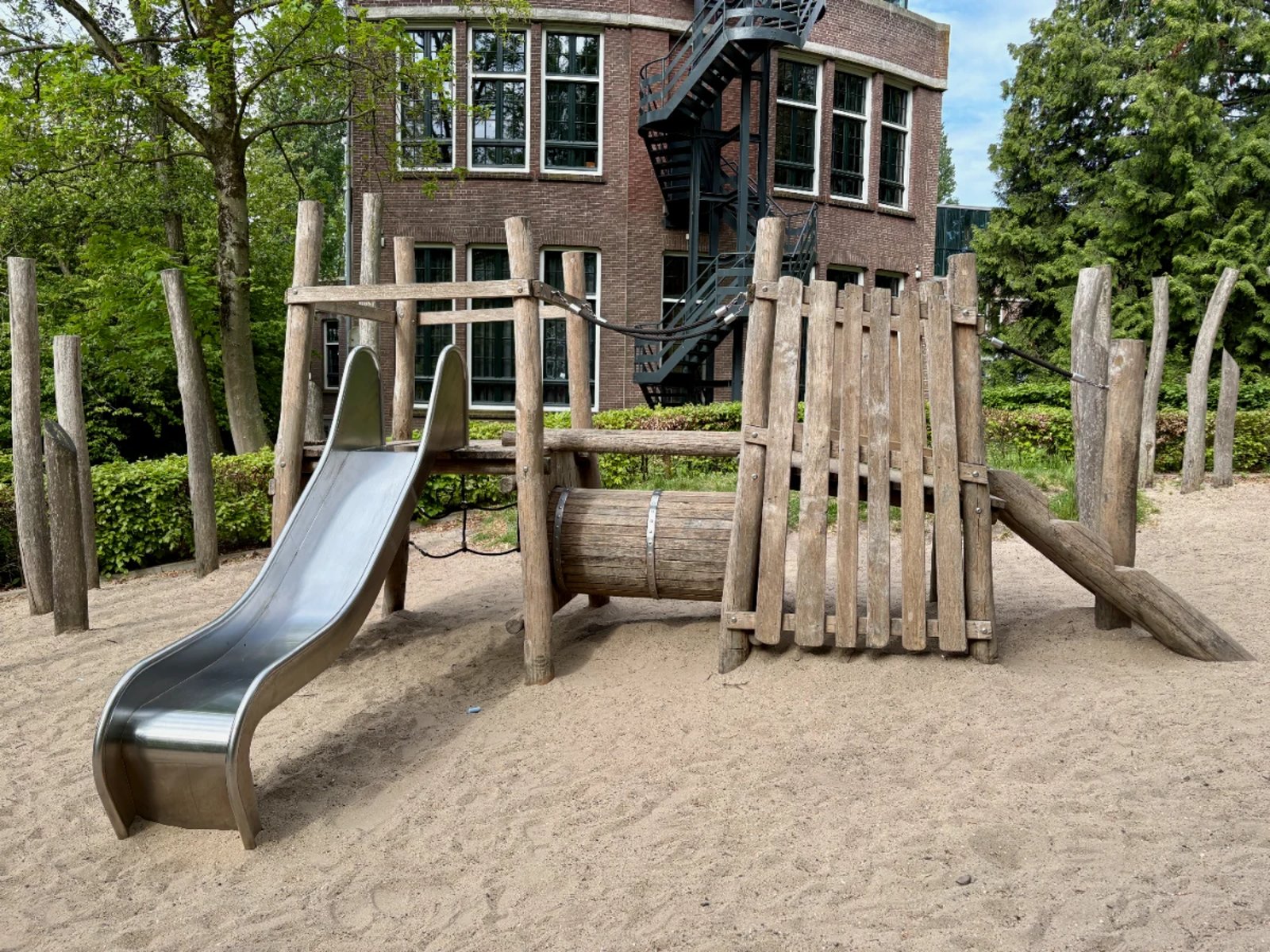 Natural wooden play structure with slide set on sand with building behind