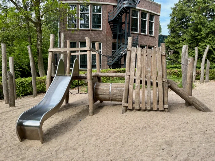 Natural wooden play structure with slide set on sand with building behind