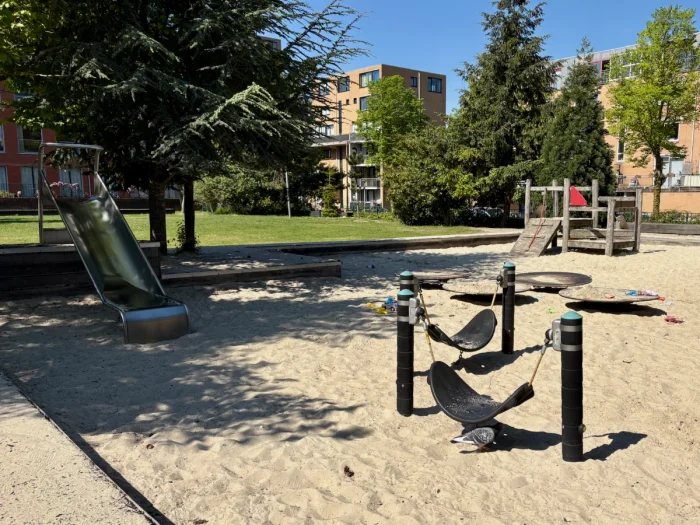Large sandbox with mini hammock swings, slide, and climbing structure in sunny park with large trees