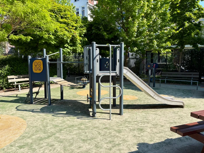 Climbing structure with slide and bridge on sunny playground with benches and trees behind 