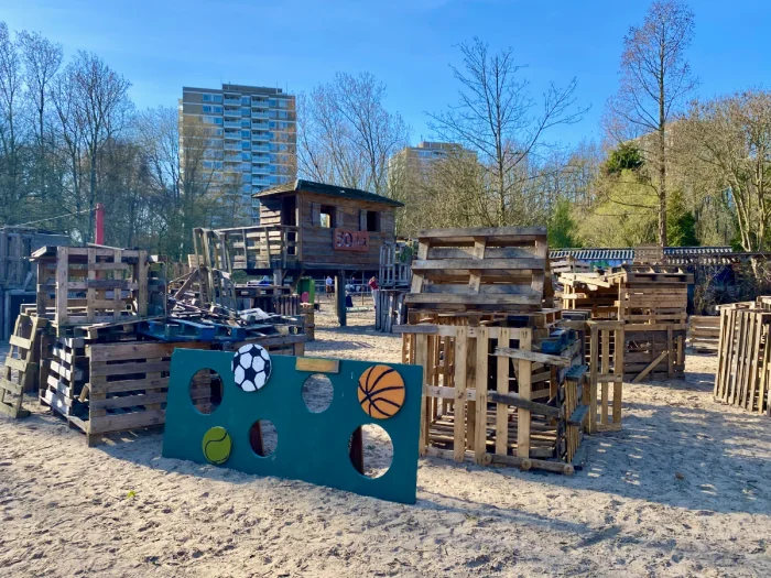 Wooden huts made out of lumber with cut-out ball game and pallets and high-rises and tree behind
