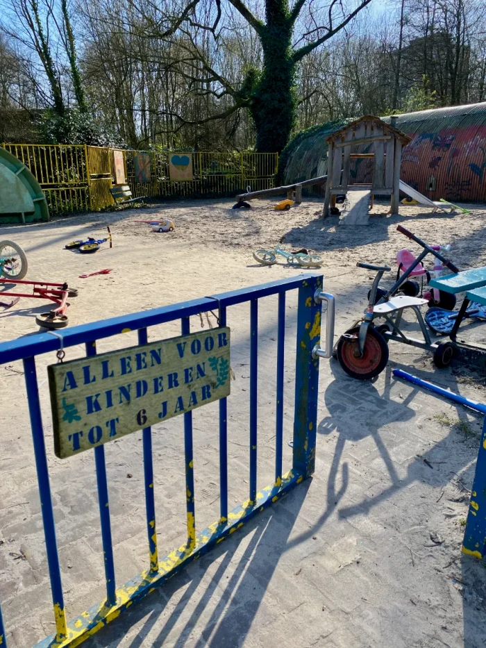 Playground with gate in the foreground and sign reading "alleen voor kinderen tot 6 jaar" and bikes and scooters inside