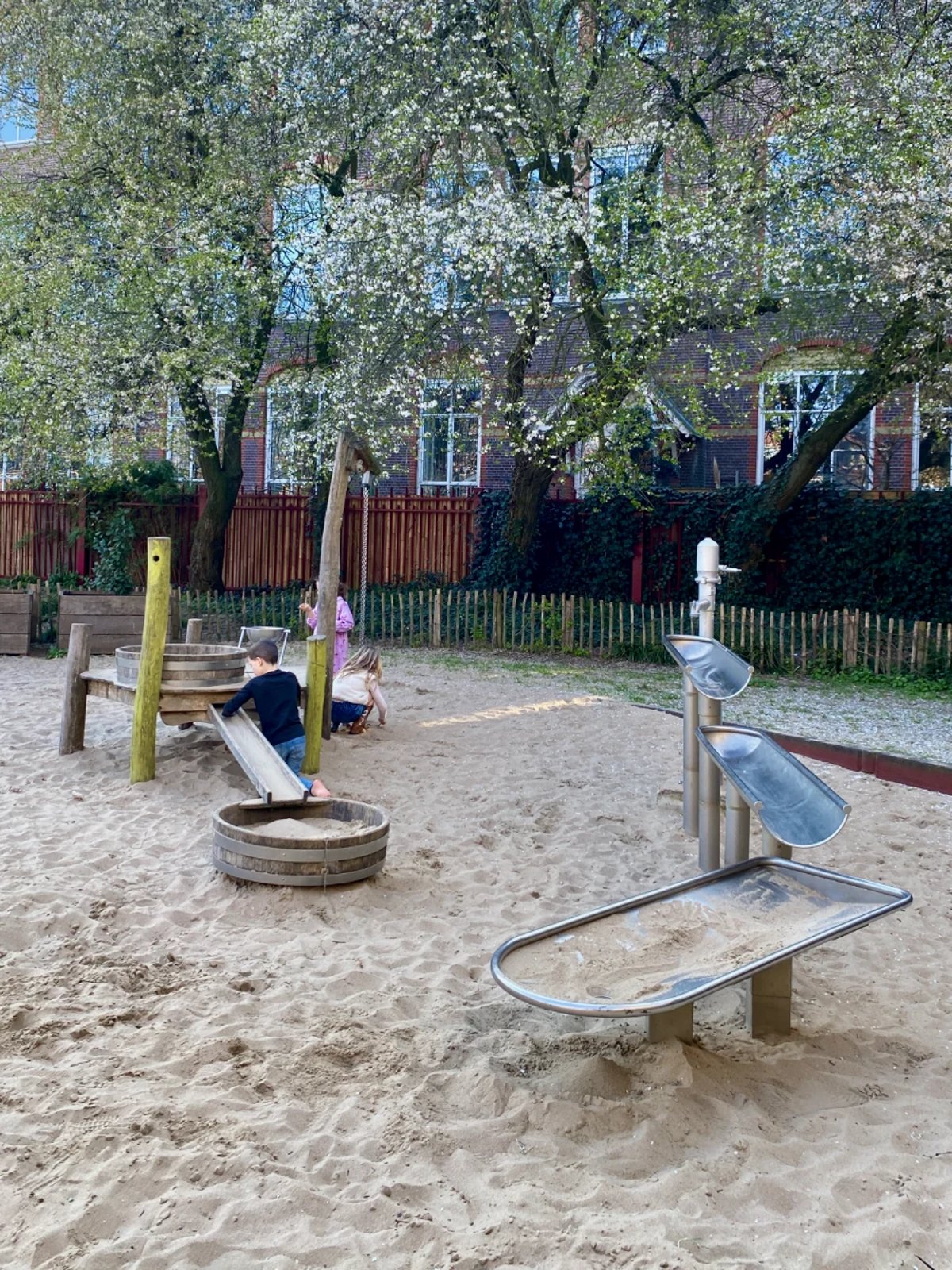Small children playing in sandbox with water channel and white-flower spring trees behind