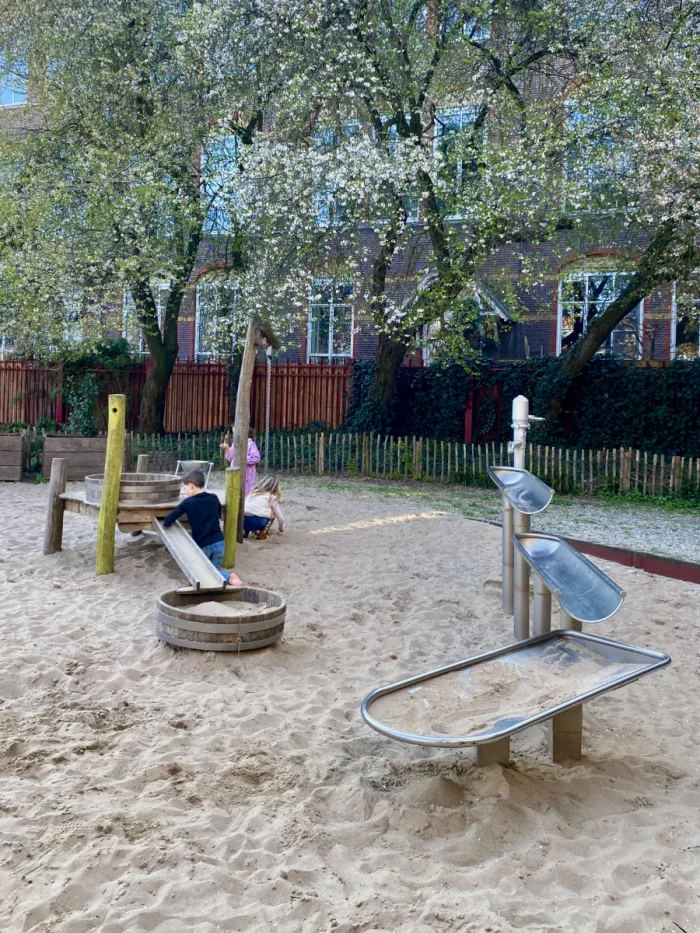 Small children playing in sandbox with water channel and white-flower spring trees behind
