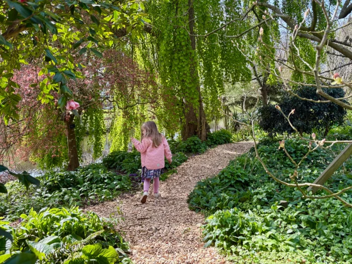 Girl in pink coat walking garden path with pink flowers and green leaves all around
