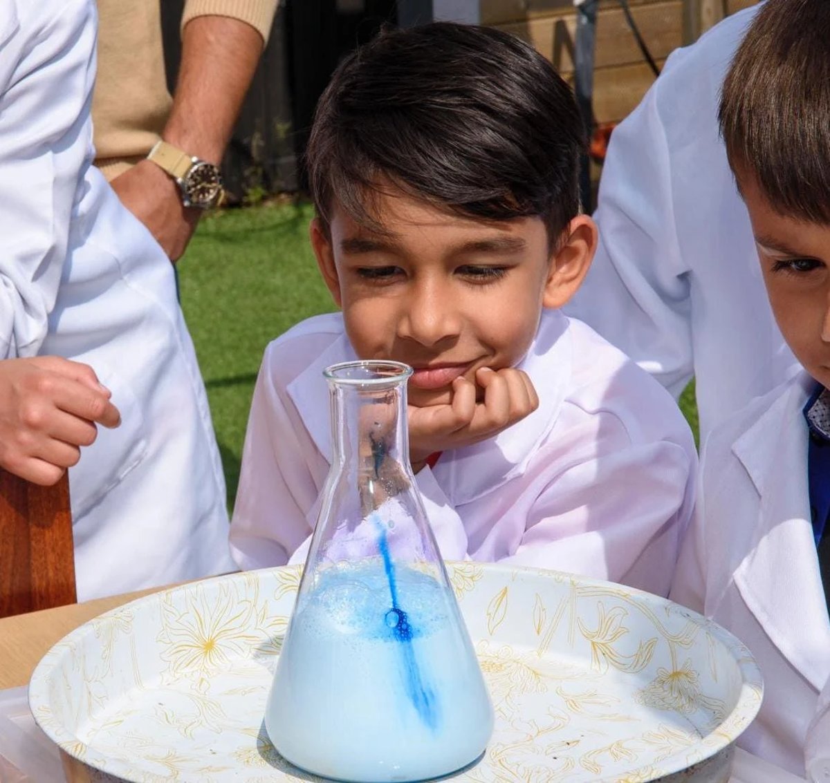 Boy with black hair and white lab coat smiles while watching a science experiment with blue liquid in a beaker