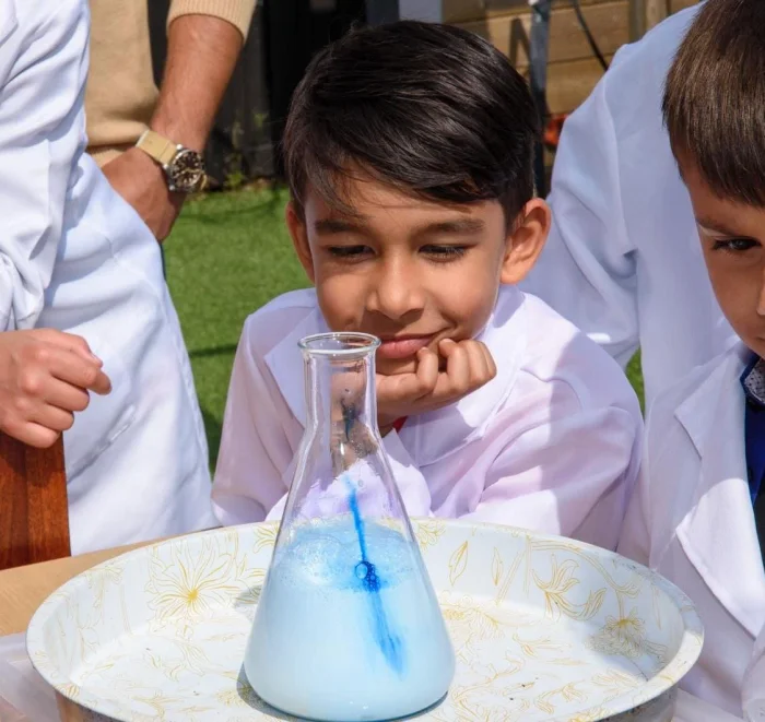 Boy with black hair and white lab coat smiles while watching a science experiment with blue liquid in a beaker