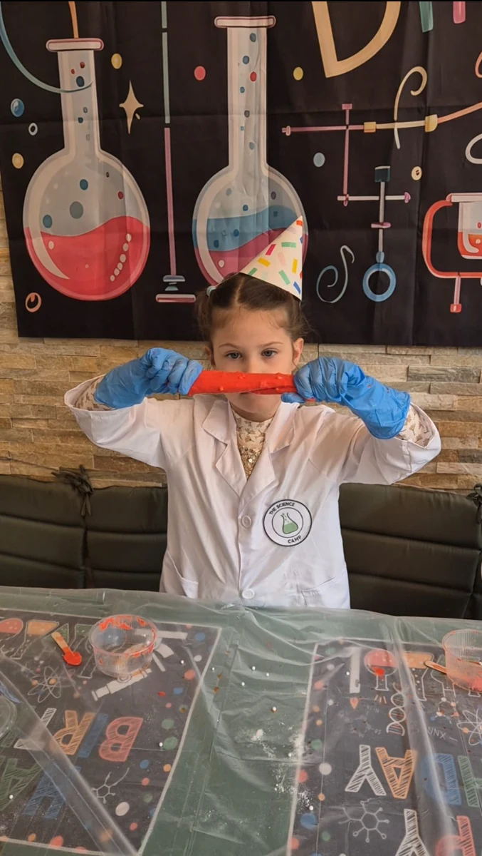 Girl with blue gloves and party hat stretches red slime in a room decorated with science illustrations