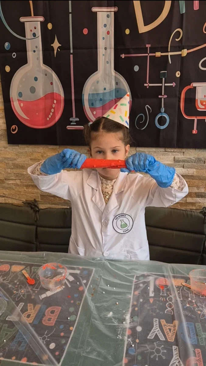 Girl with blue gloves and party hat stretches red slime in a room decorated with science illustrations